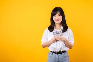 content Jeune asiatique femme d'affaires portant blanc chemise et denim jean en portant espèces, étreindre dollars argent et souriant, permanent plus de Jaune Contexte. l'amour à être riches concept. photo