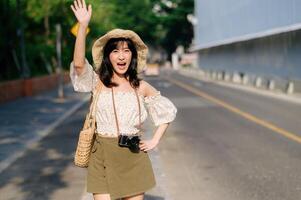 portrait de asiatique Jeune femme voyageur avec tissage chapeau et panier et une caméra agitant main à ami par le rue. périple voyage mode de vie, monde Voyage explorateur ou Asie été tourisme concept. photo