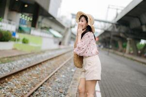 Jeune asiatique femme voyageur avec tissage panier content souriant à la recherche à une caméra à côté de train chemin de fer. périple voyage mode de vie, monde Voyage explorateur ou Asie été tourisme concept. photo