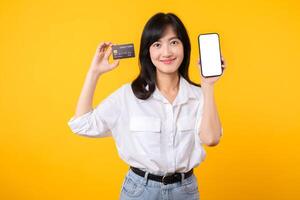 portrait asiatique Jeune femme portant blanc chemise et denim jean avec content sourire en portant crédit carte à payant achats en ligne sur téléphone intelligent isolé sur Jaune Contexte. achats en ligne concept. photo