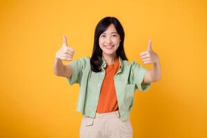 Jeune asiatique femme 30s, élégamment vêtu dans Orange chemise et vert sauteur. sa les pouces en haut geste, ensemble contre une Jaune arrière-plan, incarne le concept de affirmation. photo