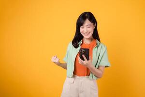 le fête de génial nouvelles avec Jeune asiatique femme 30s, élégamment vêtu dans Orange chemise et vert sauteur, élevage une poing et en portant une téléphone intelligent sur Jaune studio Contexte. mobile concept. photo