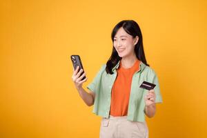 expérience le excitation de mobile achats avec vibrant Jeune asiatique femme dans sa 30s, enfiler Orange chemise et vert sauteur, en utilisant téléphone intelligent tandis que en portant crédit carte sur Jaune studio Contexte. photo