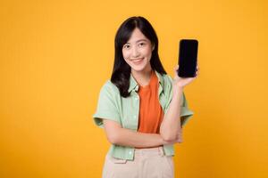 Nouveau mobile application avec de bonne humeur Jeune asiatique femme 30s, enfiler Orange chemise et vert sauteur, montrant téléphone intelligent écran sur Jaune studio Contexte. Nouveau mobile dispositif et en ligne achats concept photo