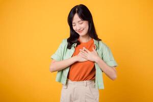 bien-être comme un asiatique femme dans sa 30s, portant une vert chemise, détient sa main sur sa poitrine contre un Orange toile de fond. soins de santé concept sur une vibrant Jaune Contexte. photo