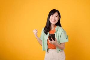 le allégresse de génial nouvelles avec une vibrant Jeune asiatique femme dans sa 30s, portant Orange chemise et vert sauteur, en portant une téléphone intelligent et élevage une poing sur Jaune studio Contexte. mobile concept. photo