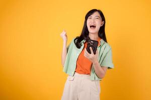 découvrir le joie de génial nouvelles avec une de bonne humeur Jeune asiatique femme dans sa 30s, portant Orange chemise et vert sauteur, en utilisant téléphone intelligent avec poing en haut geste sur Jaune Contexte. mobile concept. photo