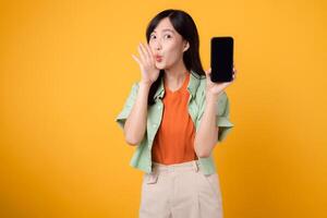 de bonne humeur Jeune asiatique femme 30s, enfiler Orange chemise et vert sauteur, mettant en valeur téléphone intelligent écran avec une crier bouche sur Jaune studio Contexte. Nouveau mobile dispositif et en ligne achats concept. photo