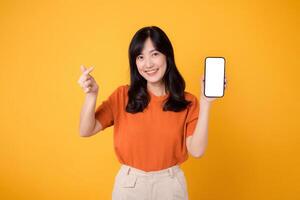 de bonne humeur Jeune asiatique femme dans sa 30s, portant un Orange chemise, montrant mini cœur main geste et téléphone intelligent Vide afficher écran sur Jaune Contexte. Nouveau application La technologie concept. photo