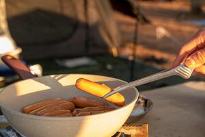 une femme main est en portant saucisses grillé sur une poêle. tandis que Aller camping à se détendre photo