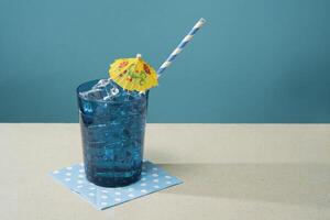 cocktail dans bleu verre rempli avec la glace avec petit Jaune parapluie et paille sur bleu serviette de table sur une blanc table photo