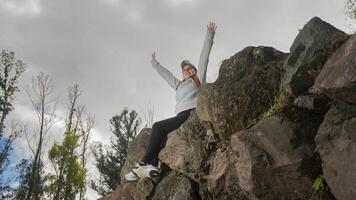 femme séance sur des pierres à le Haut de une Montagne avec sa bras élevé dans la victoire sur une nuageux journée photo