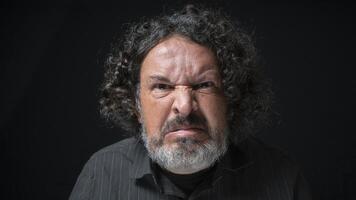 homme avec blanc barbe et noir frisé cheveux avec en colère expression, à la recherche tout droit à caméra, portant noir chemise contre noir Contexte photo