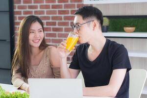 asiatique Jeune couple. Jeune homme en portant une verre de Frais Orange jus pour santé et Jeune femme à la recherche à lui tandis que séance sur une chaise dans le cuisine photo