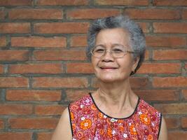 asiatique personnes âgées femme à la recherche à caméra et souriant tandis que permanent avec brique mur Contexte photo