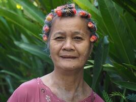 un personnes âgées asiatique femme avec cheveux rouleau, souriant et à la recherche à le caméra tandis que permanent dans une jardin photo