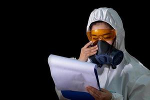 une femme tient un dossier de rapport dans des vêtements de protection chimique blancs et un masque anti-gaz avec des lunettes jaunes sur fond noir, une femme scientifique en tenue de sécurité photo