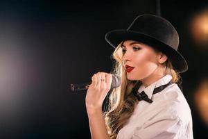 Jeune femme séduisante blonde en chemise blanche, papillon, chapeau noir avec microphone chantant sur scène photo