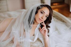 une magnifique brunette la mariée avec une tiare dans sa cheveux est en train de préparer pour le mariage dans une magnifique peignoir dans boudoir style. fermer mariage portrait de le la mariée mensonge sur le lit, photo. photo