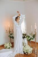 une magnifique brunette la mariée est en train de préparer pour le mariage dans une magnifique boudoir style robe parmi Frais fleurs et élégant chandeliers contre une miroir Contexte. mariage portrait, photo. photo