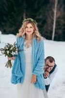 de bonne humeur portrait de une mignonne la mariée dans une blanc robe avec une bouquet de fleurs dans sa mains, enveloppé dans une bleu plaid, et le jeune marié contre le Contexte de une hiver paysage de enneigé montagnes. photo
