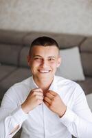 le jeune marié boutons le sien chemise par le la fenêtre. homme dans une chemise. Masculin portrait. séance photo à le la fenêtre. du marié collection. mariage journée. Beau homme jeune marié boutonnage le sien chemise dans une Hôtel chambre.