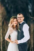 portrait de le la mariée et jeune marié contre le Contexte de une pin forêt. le la mariée dans une blanc mariage robe avec une couronne sur sa diriger, le jeune marié dans une blanc chemise et gilet. hiver mariage concept. photo