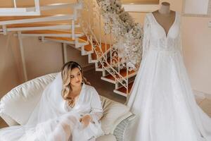 Matin de le la mariée avant le mariage. une magnifique Jeune femme avec une voile et une magnifique coiffure dans une blanc peignoir près sa mariage robe sur une mannequin. professionnel se maquiller. dernier les préparatifs. photo