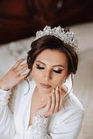 une magnifique brunette la mariée avec une tiare dans sa cheveux est avoir prêt pour le mariage dans une magnifique peignoir dans boudoir style. fermer mariage portrait, photo. photo