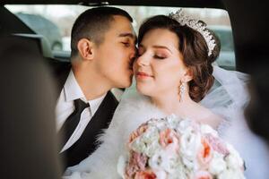 portrait de incroyablement magnifique les mariées dans le fenêtre de un coûteux auto. la mariée avec une bouquet de fleurs. le jeune marié baisers le la mariée. photo