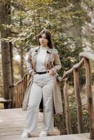 verticale photo. magnifique insouciant Jeune adolescent fille dans marron manteau et blanc jeans et blanc chandail. portrait de une magnifique fille dans une marron imperméable contre le Contexte de le forêt et la nature. photo