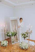 une magnifique brunette la mariée est en train de préparer pour le mariage dans une magnifique boudoir style robe parmi Frais fleurs et élégant chandeliers contre une miroir Contexte. mariage portrait, photo. photo