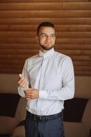 le jeune marié boutons le sien chemise par le la fenêtre. homme dans une chemise. Masculin portrait. séance photo à le la fenêtre. du marié collection. mariage journée. Beau homme jeune marié boutonnage le sien chemise dans une Hôtel chambre.