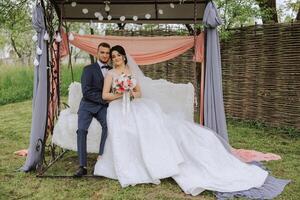 une magnifique Jeune la mariée dans une printemps parc embrasse sa jeune marié tandis que séance sur une balançoire dans le jardin. magnifique mariage blanc robe avec une longue train. des promenades dans le parc. une content et aimant couple. photo