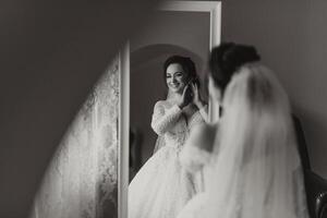 le la mariée dans une mariage robe pose dans sa chambre, à la recherche dans le miroir. noir et blanc photo. Matin de le la mariée. préparation. mariage la photographie photo