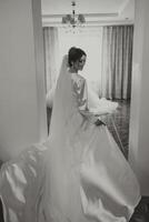 une magnifique brunette la mariée est avoir prêt pour le mariage dans une magnifique boudoir style tenue en marchant à sa mariage robe par le la fenêtre. noir et blanc photo. photo