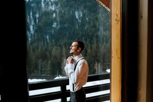 Jeune homme portant bretelles, boutonnage le sien pie tandis que permanent sur une balcon avec une vue de enneigé montagnes. du repos dans le montagnes. Montagne air. mariage dans hiver photo