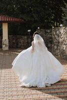 le la mariée dans une magnifique robe et voile des promenades vers le bas le pierre chemin avec sa épaules tourné. magnifique robe avec longue manches, ouvert bousiller. été mariage photo