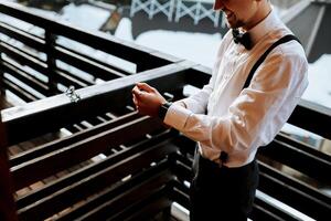 une Jeune homme portant bretelles, une blanc chemise et une noir arc attacher met sur le sien montre. une mariage dans le montagnes dans hiver photo