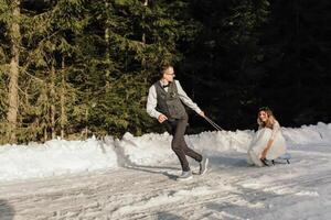 le la mariée et jeune marié balade une traîneau sur une neigeux route dans l'hiver. hiver mariage sur le rue dans le forêt. le hiver vacances. content couple luge dans neigeux hiver temps photo