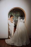 une content la mariée est en train de préparer pour sa luxueux mariage dans une Hôtel chambre, avec une mariage robe sur une mannequin proche. portrait de une femme avec à la mode cheveux, maquillage et une sourire dans une pansement robe. photo