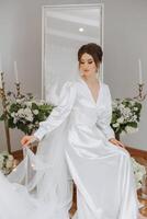 une magnifique brunette la mariée est avoir prêt pour le mariage dans une magnifique boudoir style tenue séance sur une chaise parmi Frais fleurs et élégant chandeliers. mariage portrait, photo. photo