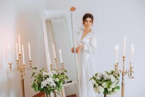 une magnifique brunette la mariée est en train de préparer pour le mariage dans une magnifique boudoir style robe parmi Frais fleurs et élégant chandeliers contre une miroir Contexte. mariage portrait, photo. photo