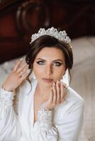 une magnifique brunette la mariée avec une tiare dans sa cheveux est avoir prêt pour le mariage dans une magnifique peignoir dans boudoir style. fermer mariage portrait, photo. photo