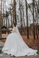 le la mariée est portant une blanc volumineux robe avec une longue former, en portant une bouquet et posant tandis que en marchant dans le forêt. hiver mariage. photo