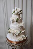 une grand luxueux à plusieurs niveaux mariage gâteau est décoré avec Frais blanc Rose fleurs dans le banquet salle. mariage dessert en dessous de le soir lumière. mariage décor. photo