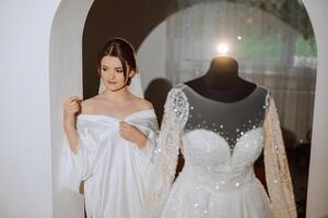 une content la mariée est en train de préparer pour sa luxueux mariage dans une Hôtel chambre, avec une mariage robe sur une mannequin proche. portrait de une femme avec à la mode cheveux, maquillage et une sourire dans une pansement robe. photo