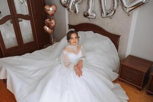 une luxueux la mariée avec une magnifique coiffure et une magnifique robe est avoir prêt pour le mariage la cérémonie dans le Matin. Matin photo de le la mariée à Accueil ou dans une Hôtel chambre. professionnel se maquiller.