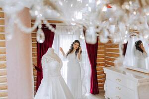 une content la mariée est en train de préparer pour sa luxueux mariage dans une Hôtel chambre, avec une mariage robe sur une mannequin proche. portrait de une femme avec à la mode cheveux, maquillage et une sourire dans une pansement robe. photo