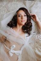 une magnifique brunette la mariée avec une tiare dans sa cheveux est en train de préparer pour le mariage dans une magnifique peignoir dans boudoir style. fermer mariage portrait de le la mariée mensonge sur le lit, photo de au-dessus de.